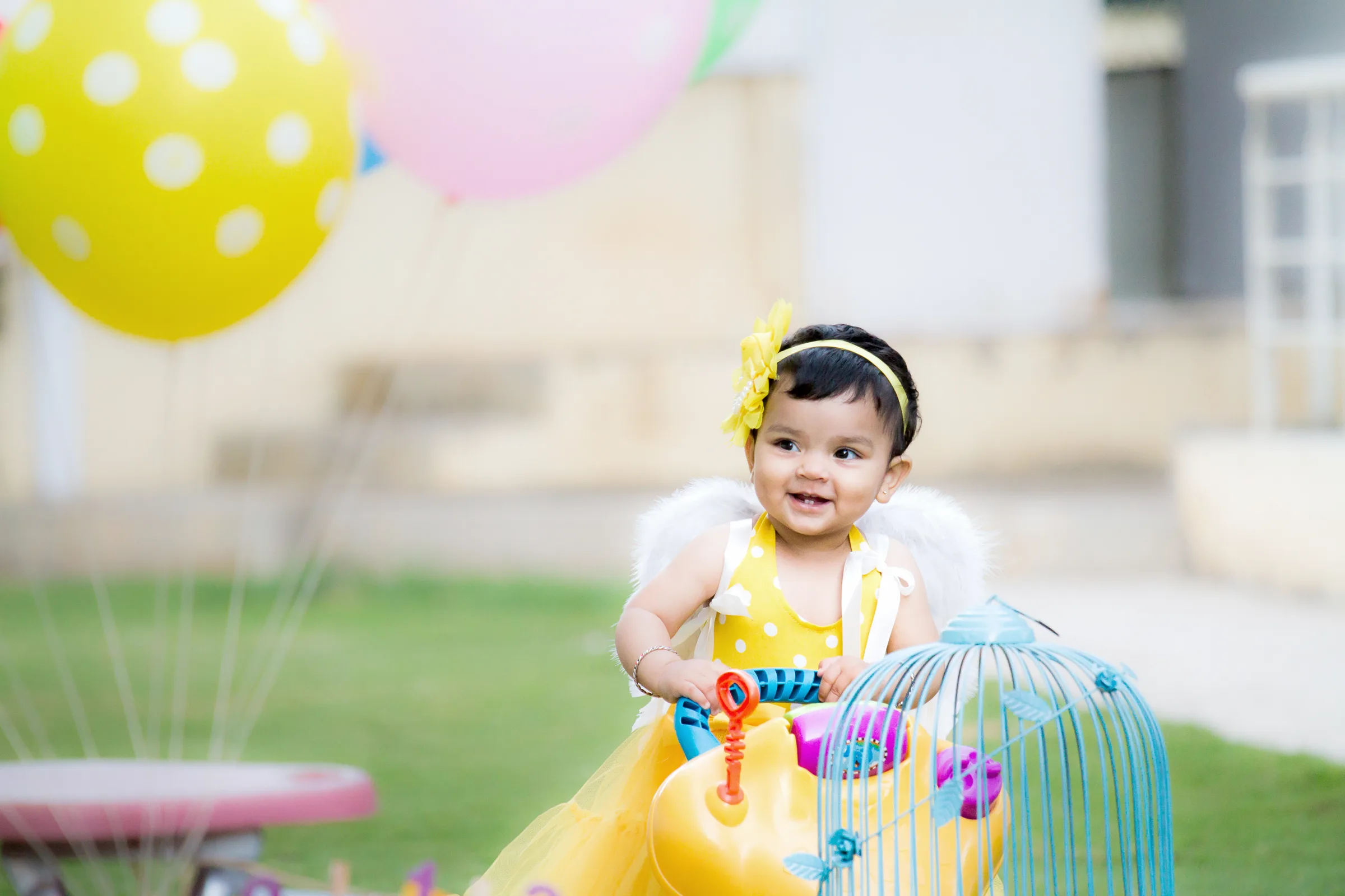 Baby girl in yellow with angel wings playing beside a toy birdcage and balloons outdoors, kids photography by Vivek Pankaj