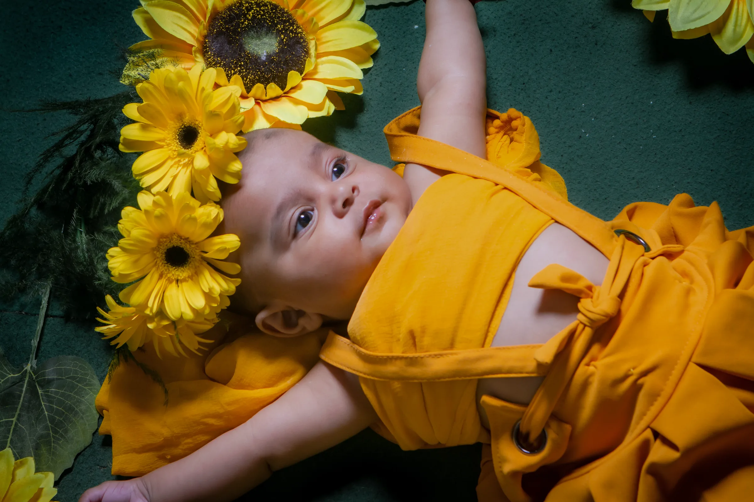 Baby in a mustard romper lying among sunflowers and gerberas, looking up with wide eyes, kids photography by Vivek Pankaj