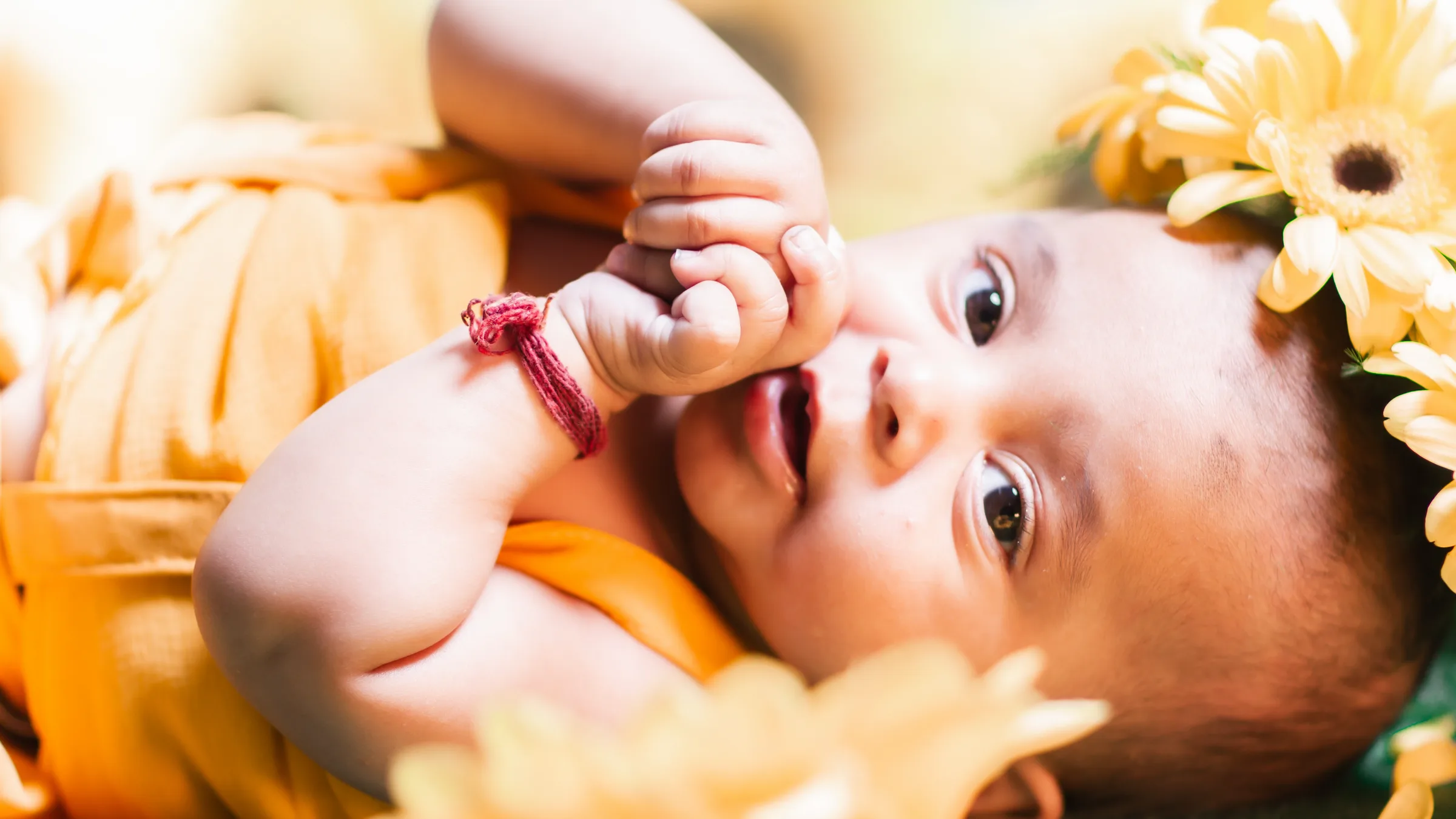 Baby in mustard with a red thread bracelet sucking on her fist among yellow gerberas, kids photography by Vivek Pankaj