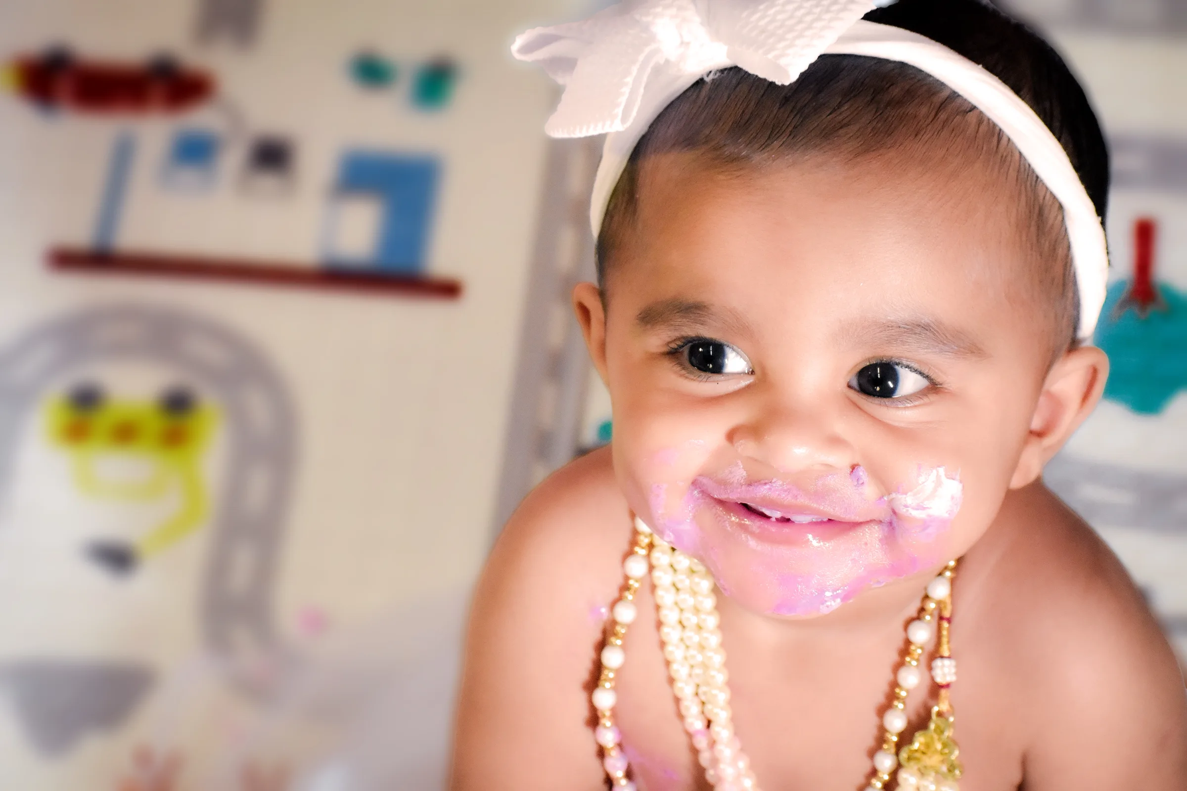 Baby with a white bow headband and cake on her face, smiling during a first birthday shoot by Vivek Pankaj