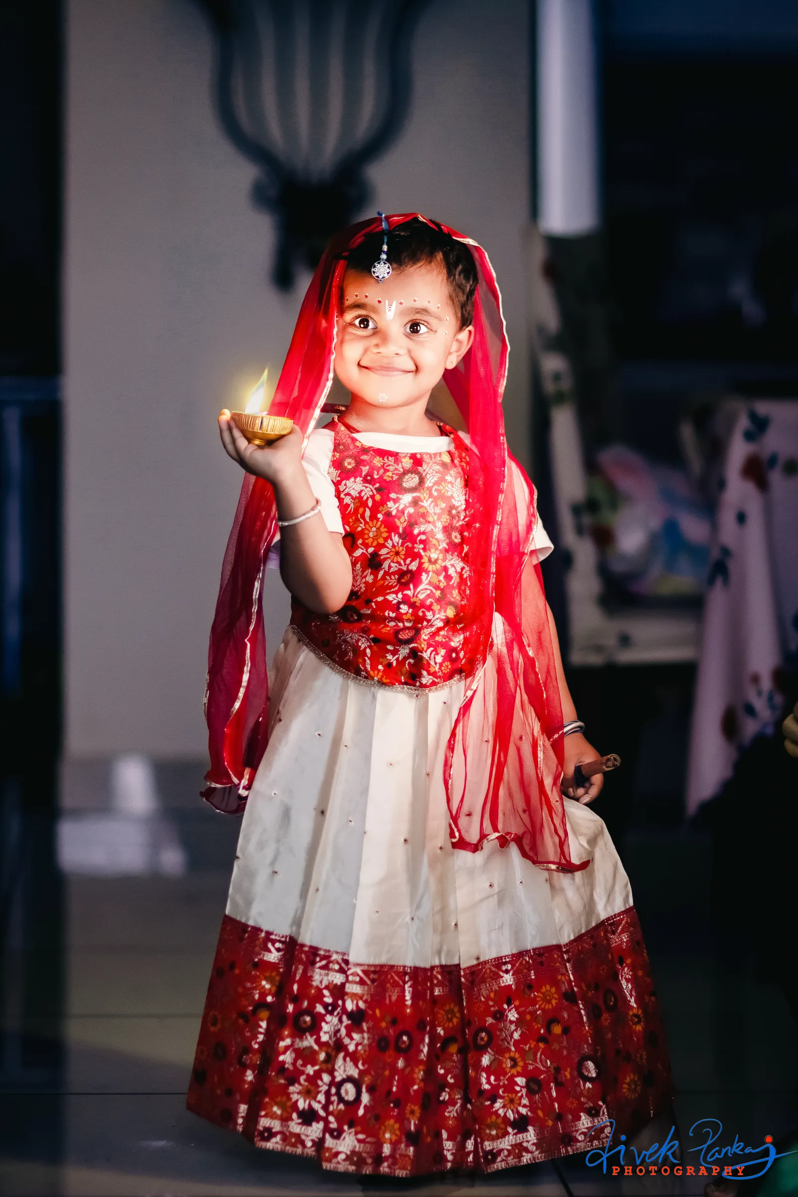 Little girl in a red and white lehenga holding a lit diya, festive kids portrait by Vivek Pankaj