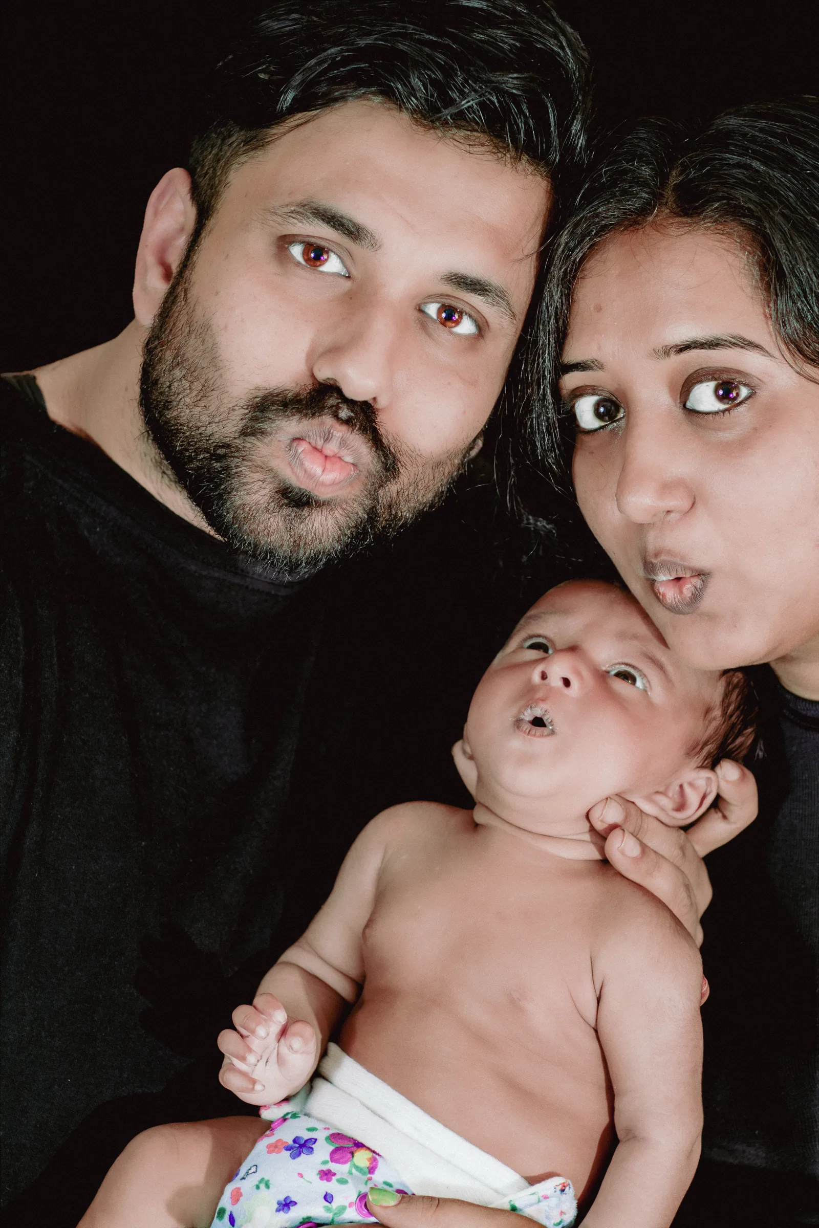 Playful family portrait of parents pulling surprised pouting faces with their wide-eyed newborn copying the expression, black backdrop, newborn and family photography by Vivek Pankaj