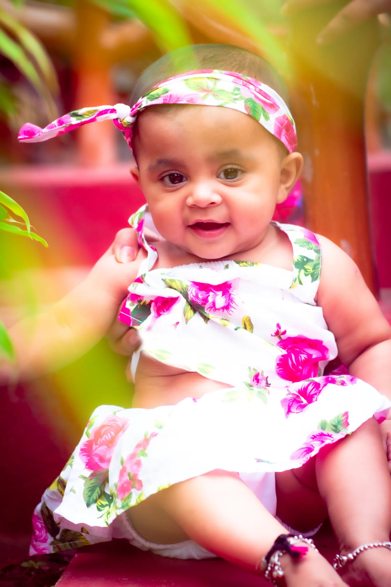 Smiling baby girl in a pink floral dress and matching headband sitting outdoors in soft sunlight, kids photography by Vivek Pankaj