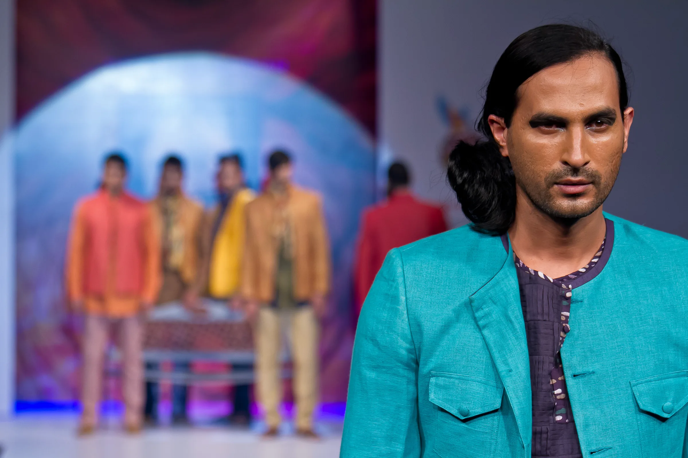 Male model in a turquoise jacket with a line of menswear in bright colours behind him at Bangalore Fashion Week, photographed by Vivek Pankaj