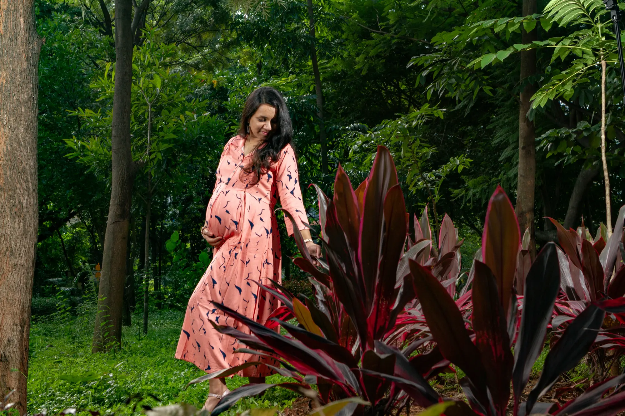 Expectant mother in a pink print dress walking past red foliage in a green forest, maternity photography by Vivek Pankaj
