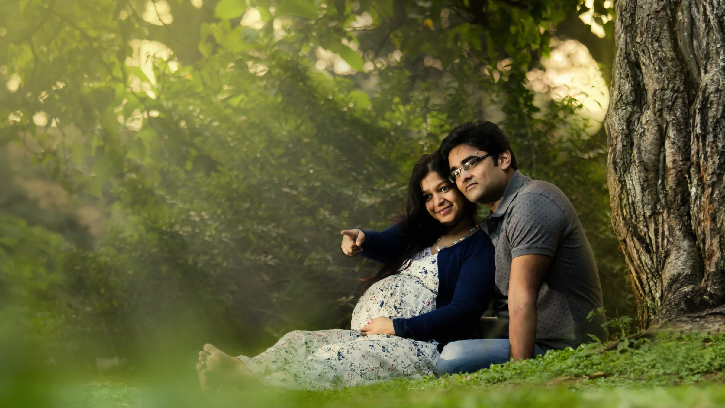 Expecting couple relaxing on the grass in sunlit woods, pointing into the distance, maternity shoot by Vivek Pankaj