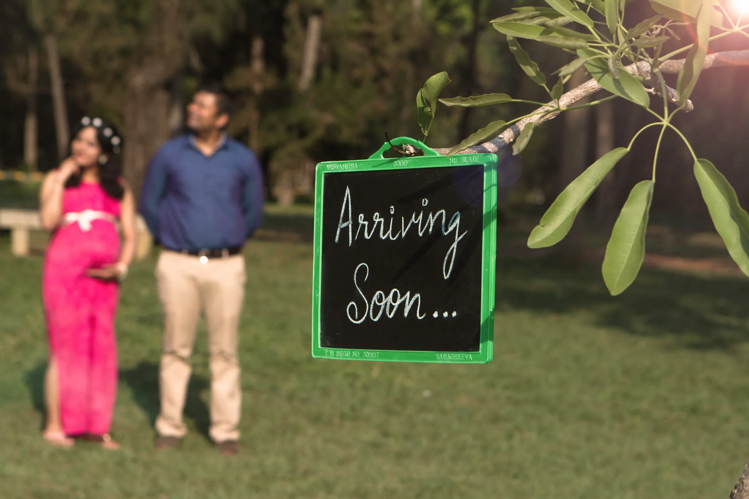 Expecting couple standing beside an 'Arriving Soon' chalkboard sign on a lawn, maternity photoshoot by Vivek Pankaj