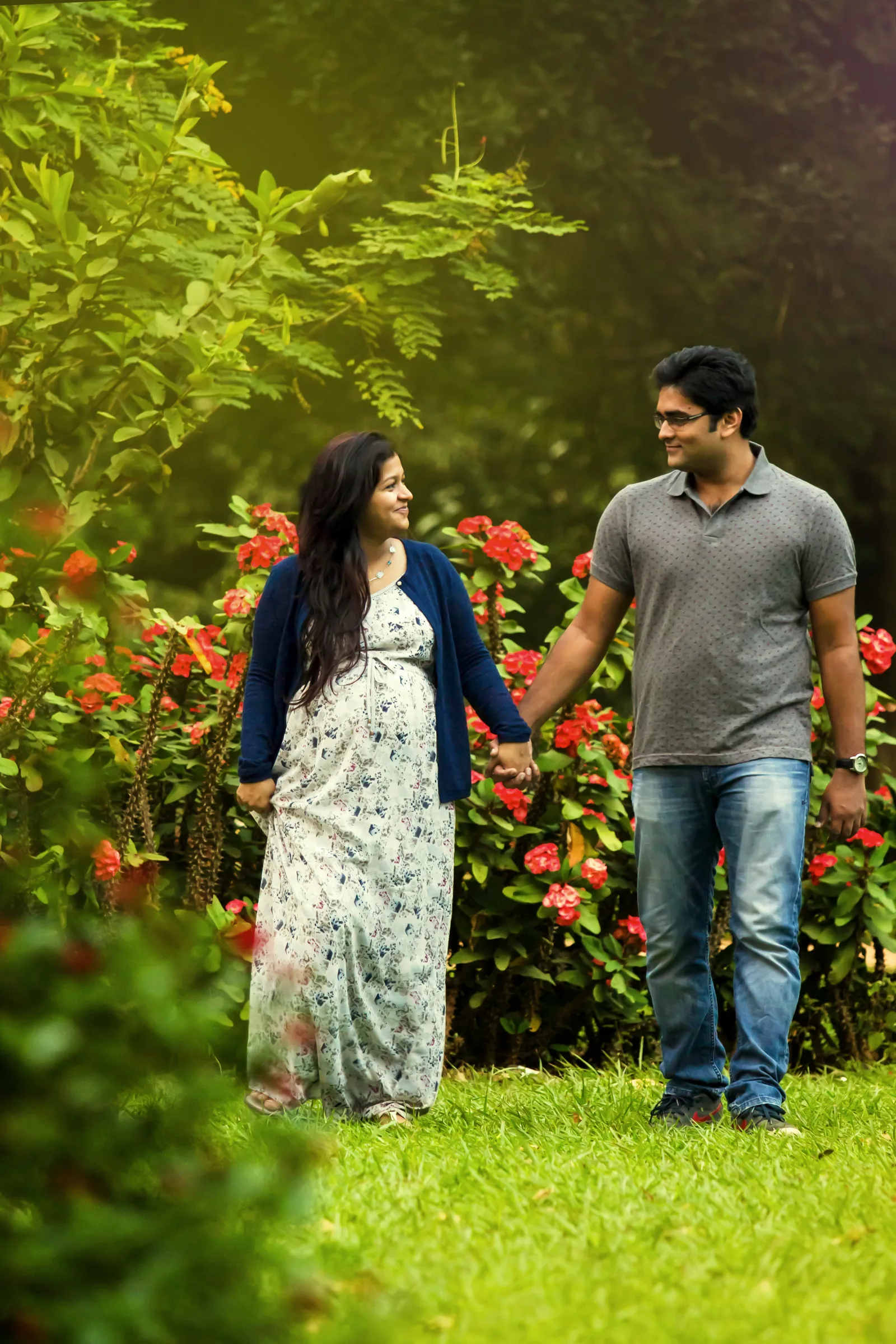 Expecting couple walking hand in hand on grass past red flowers, maternity photography by Vivek Pankaj