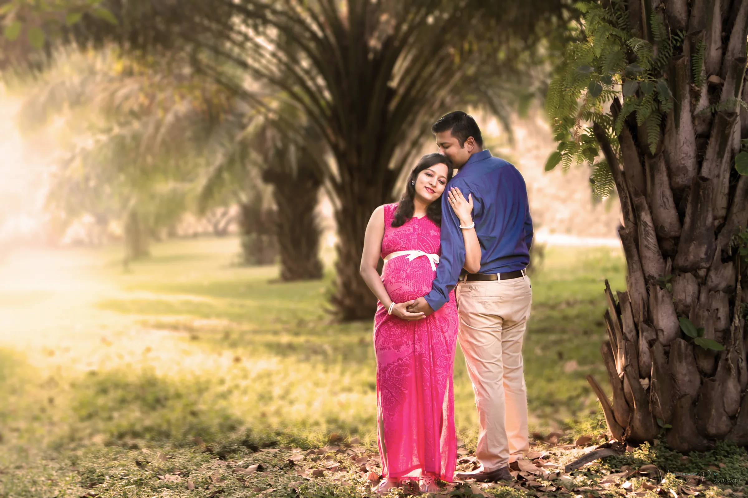 Husband embracing his pregnant wife in a pink gown in a palm grove, maternity photoshoot by Vivek Pankaj