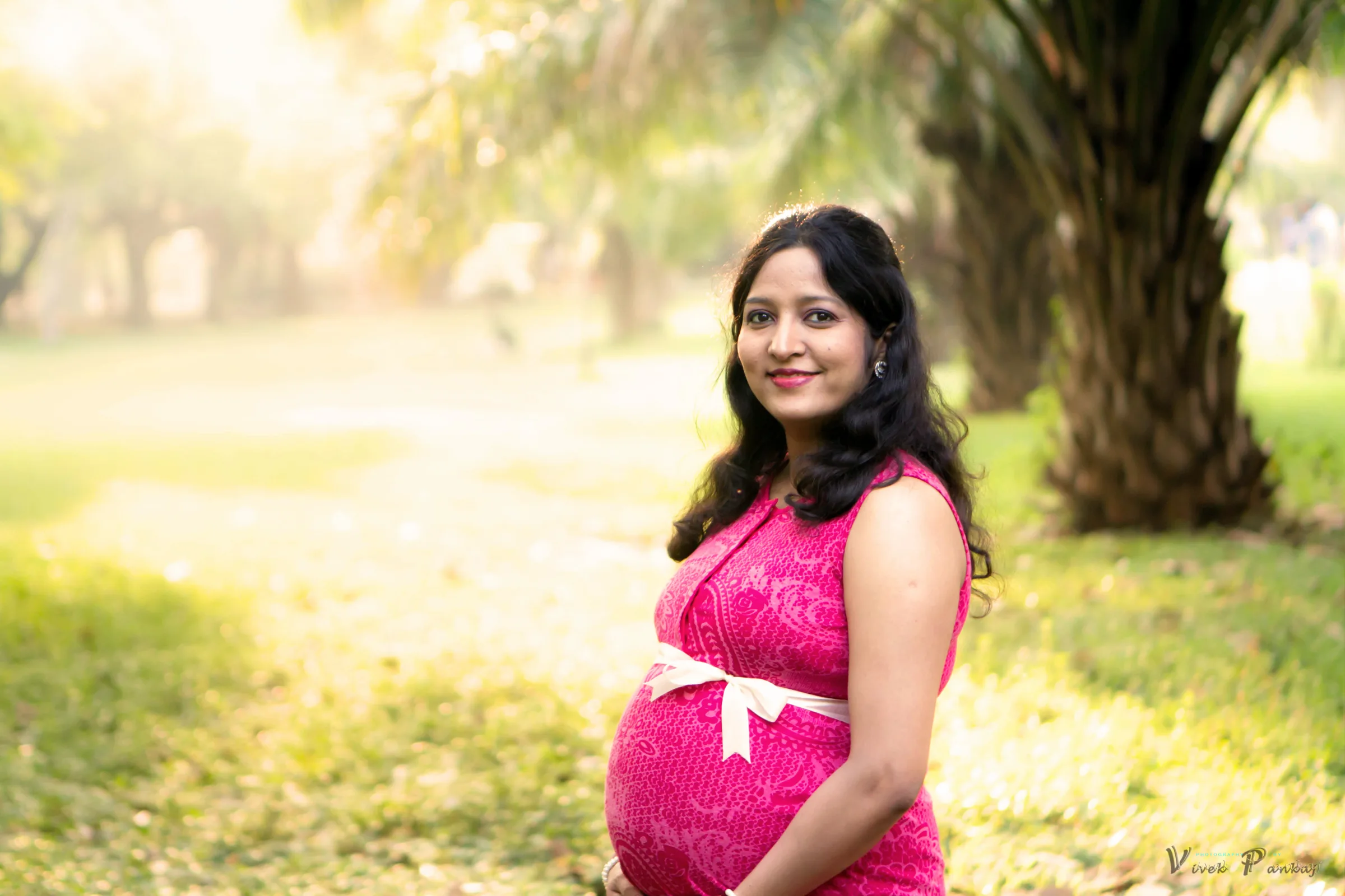 Mother-to-be in a pink dress cradling her bump in soft golden light among palm trees, maternity portrait by Vivek Pankaj