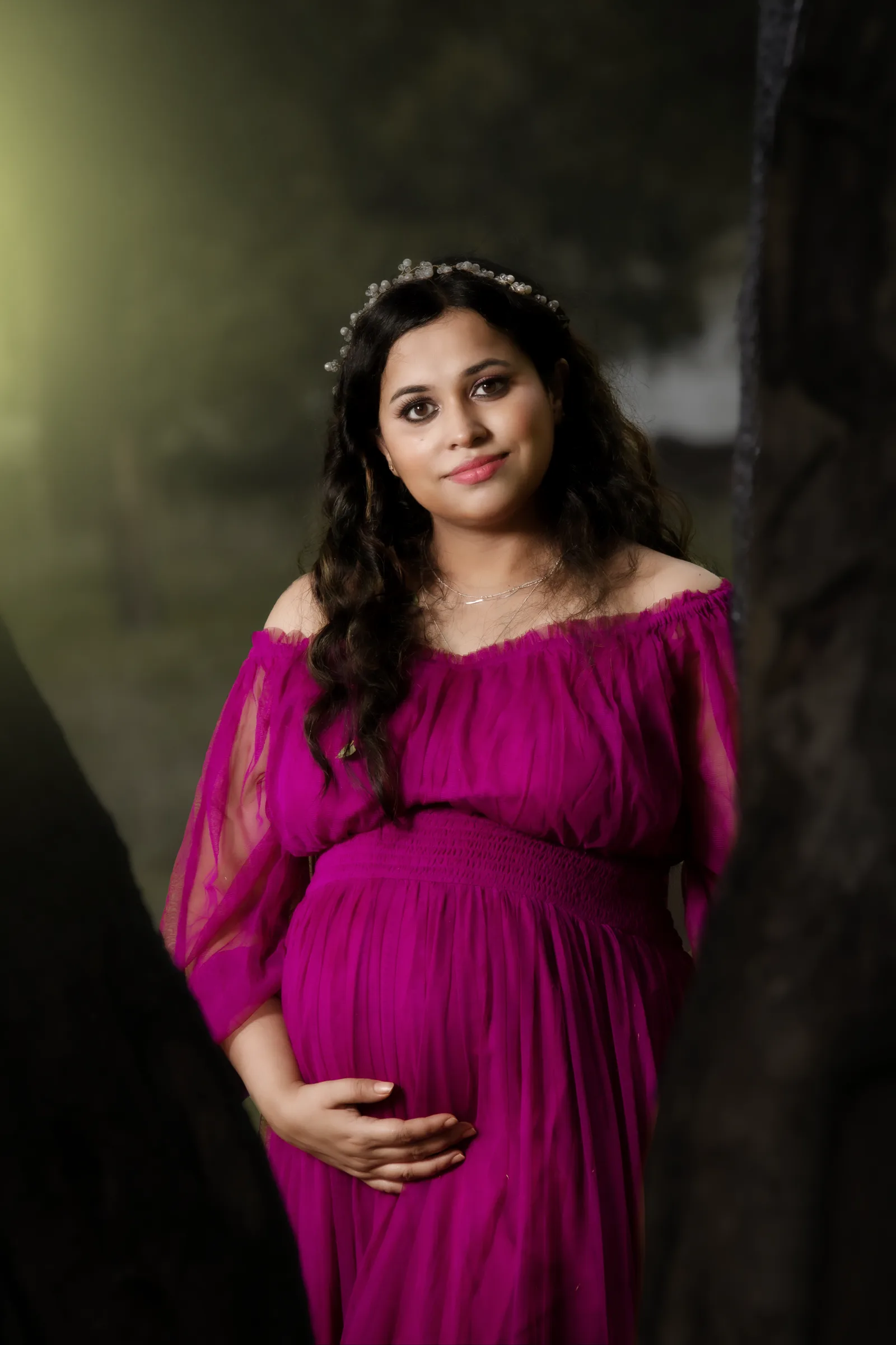 Mother-to-be in an off-shoulder magenta gown and flower crown against a tree, maternity portrait by Vivek Pankaj