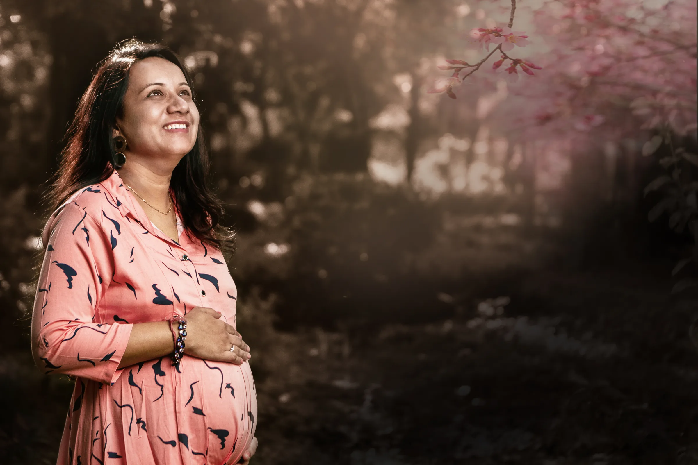Smiling expectant mother in a pink dress looking up in a misty forest, maternity portrait by Vivek Pankaj