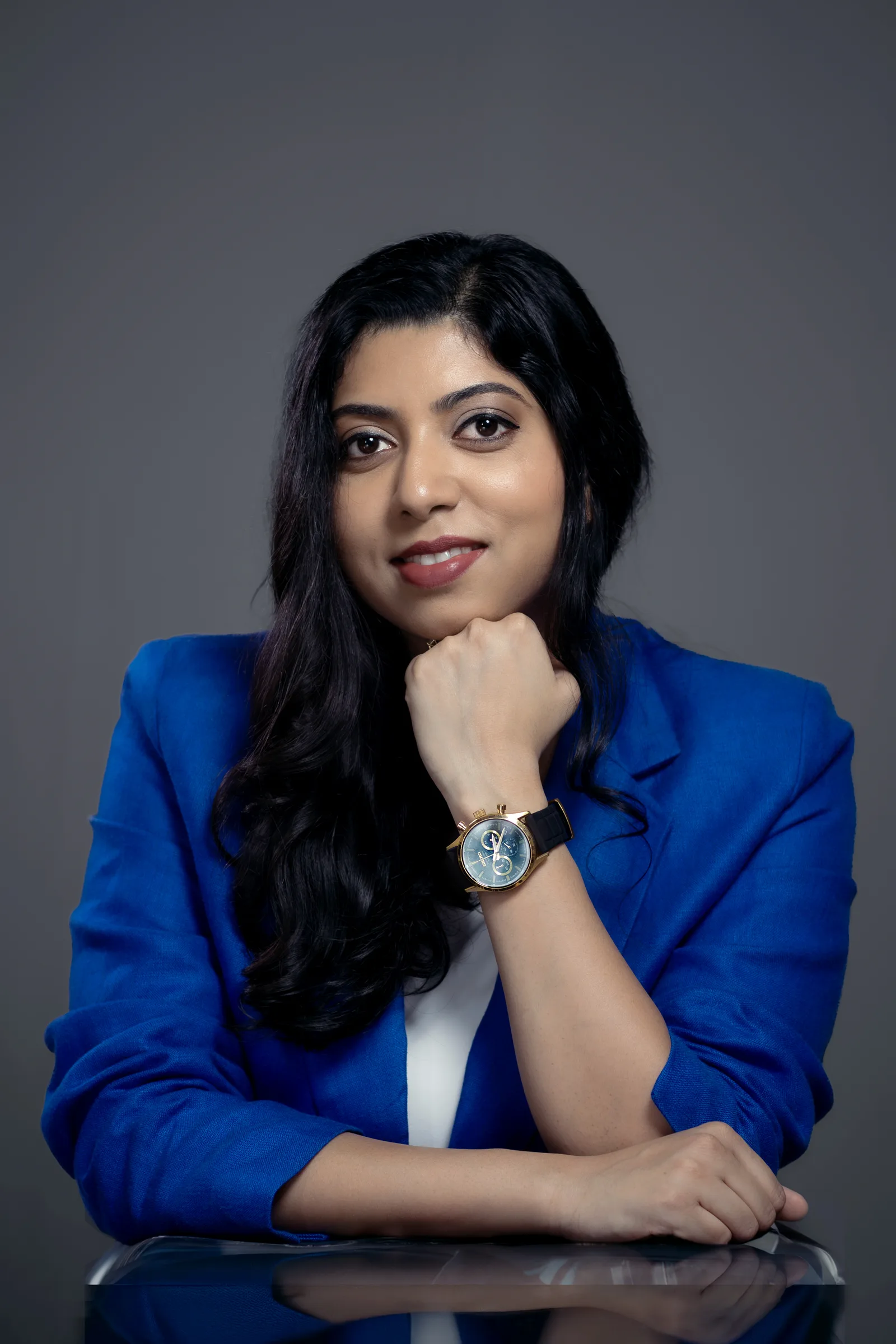 Professional studio portrait of a smiling woman in a royal blue blazer resting her chin on her hand at a glass table, grey backdrop, corporate headshot by Vivek Pankaj