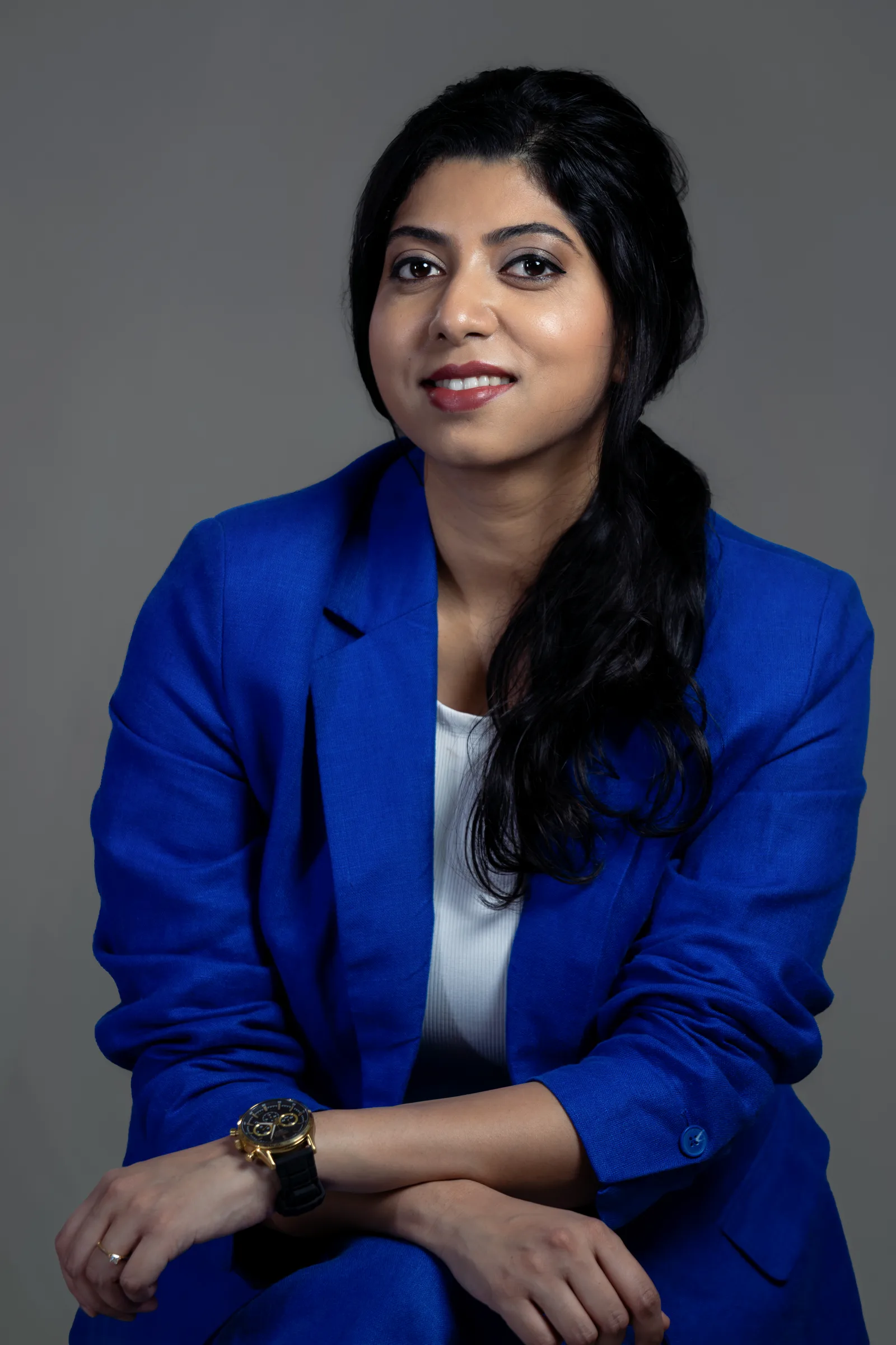 Seated studio portrait of a smiling woman in a royal blue blazer and white top with a gold watch, arms resting on her knee, grey backdrop, professional portrait by Vivek Pankaj