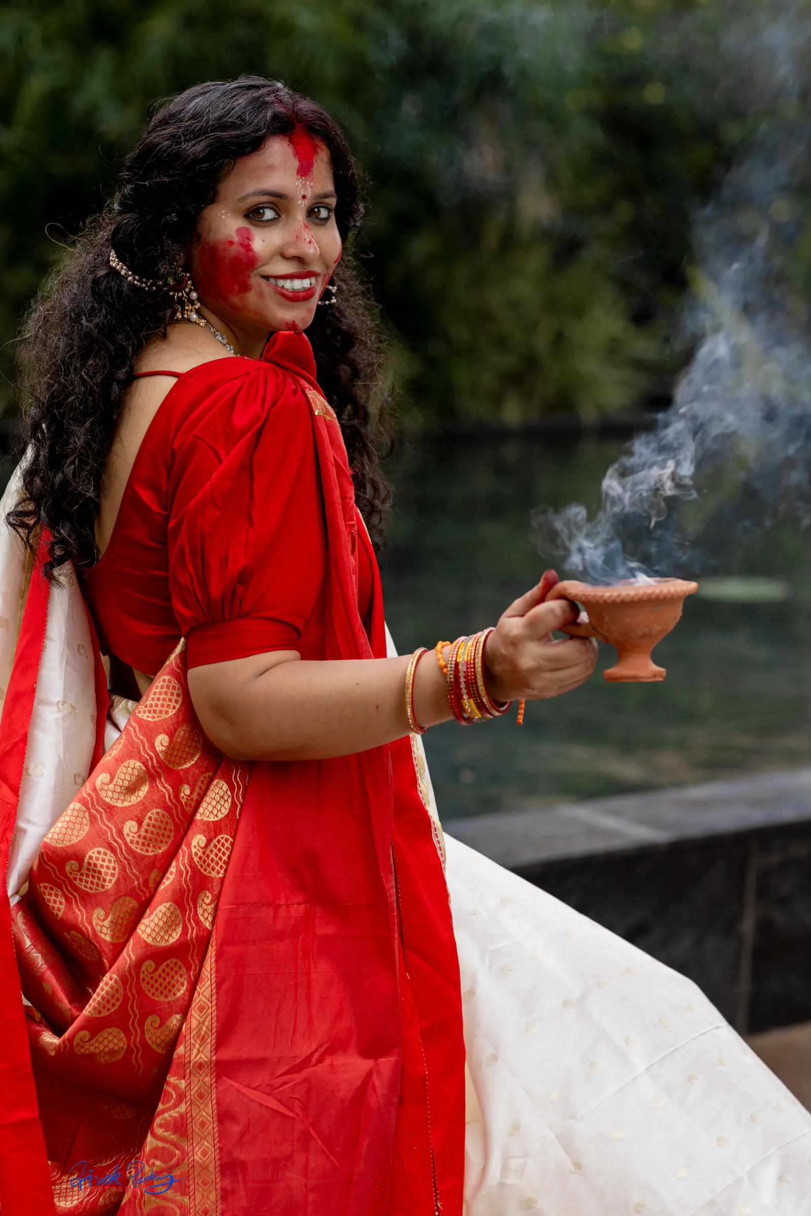 Smiling woman in a red and white Bengali saree with sindoor on her face, holding a smoking clay dhunuchi, festive portrait, photography by Vivek Pankaj