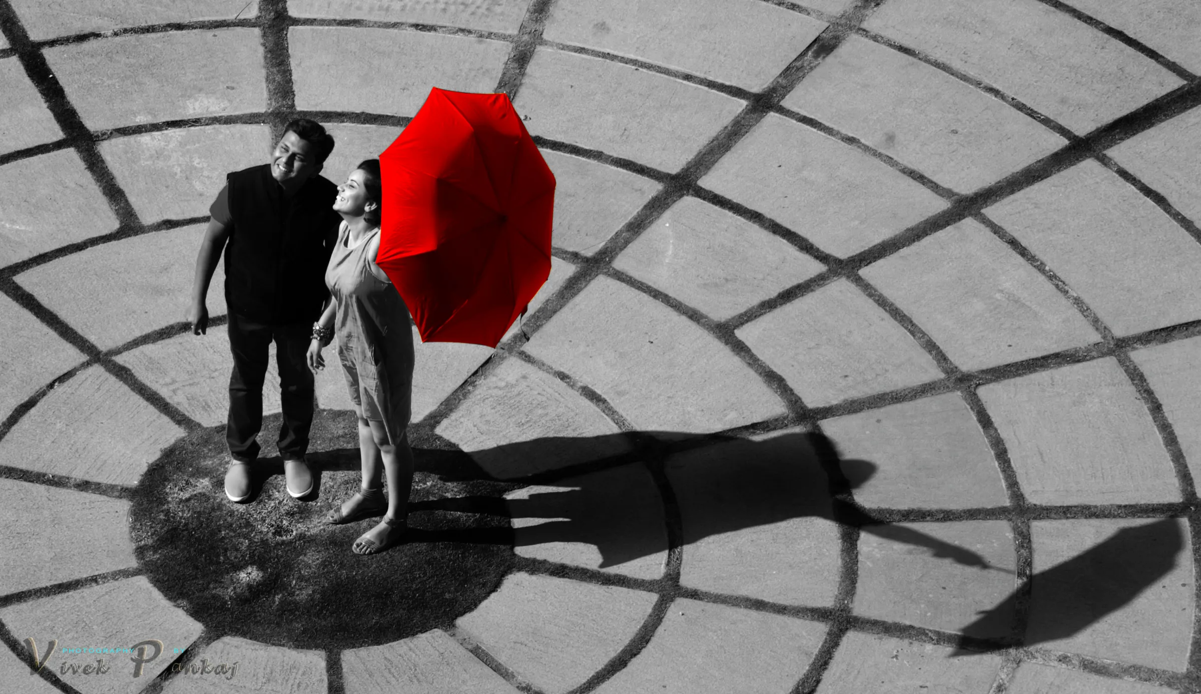 Black and white top-down frame of a couple under a single red umbrella on a patterned courtyard, pre-wedding photography by Vivek Pankaj