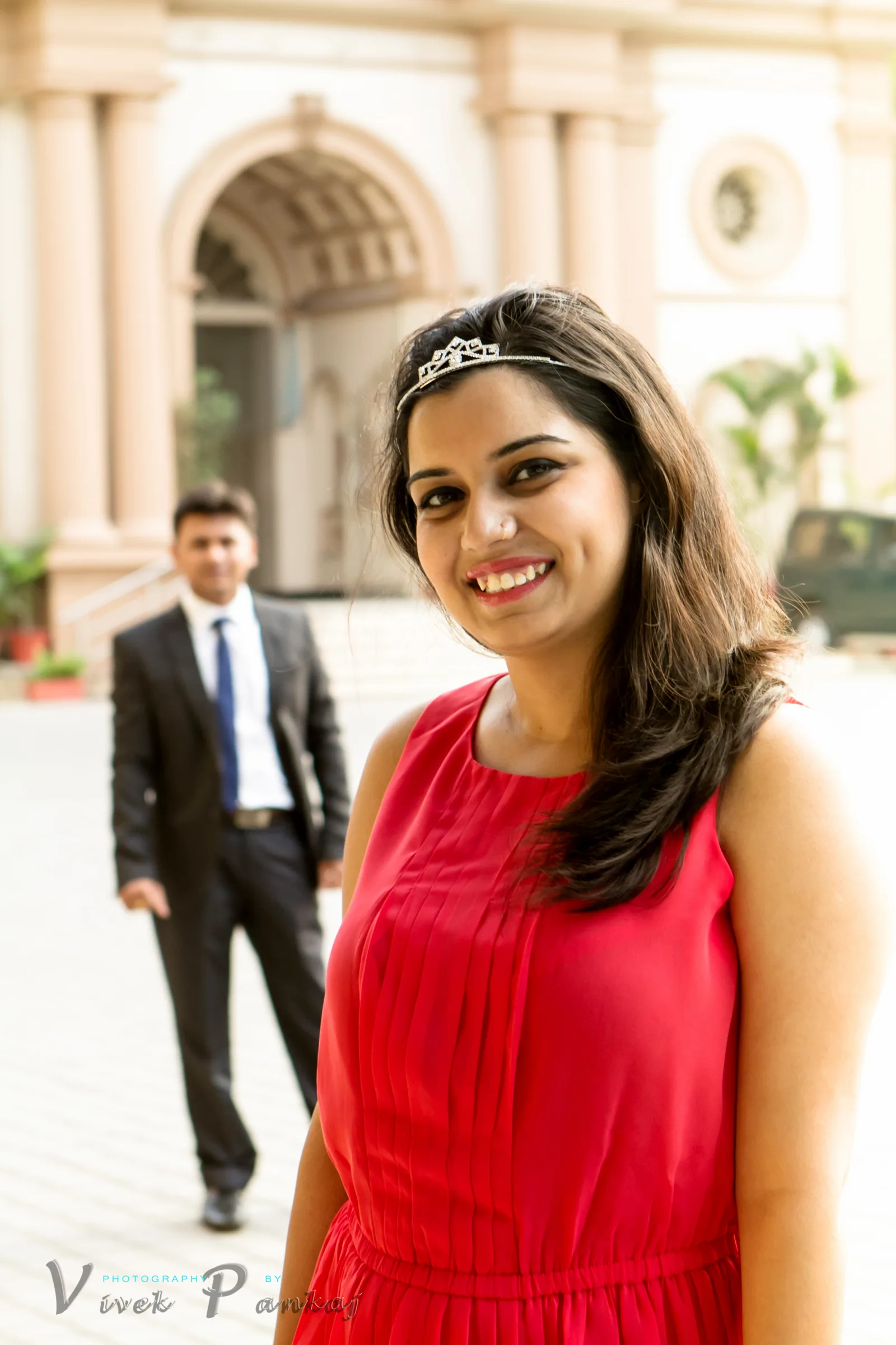 Smiling bride-to-be in a red dress and tiara with her partner in a suit behind her, pre-wedding portrait by Vivek Pankaj