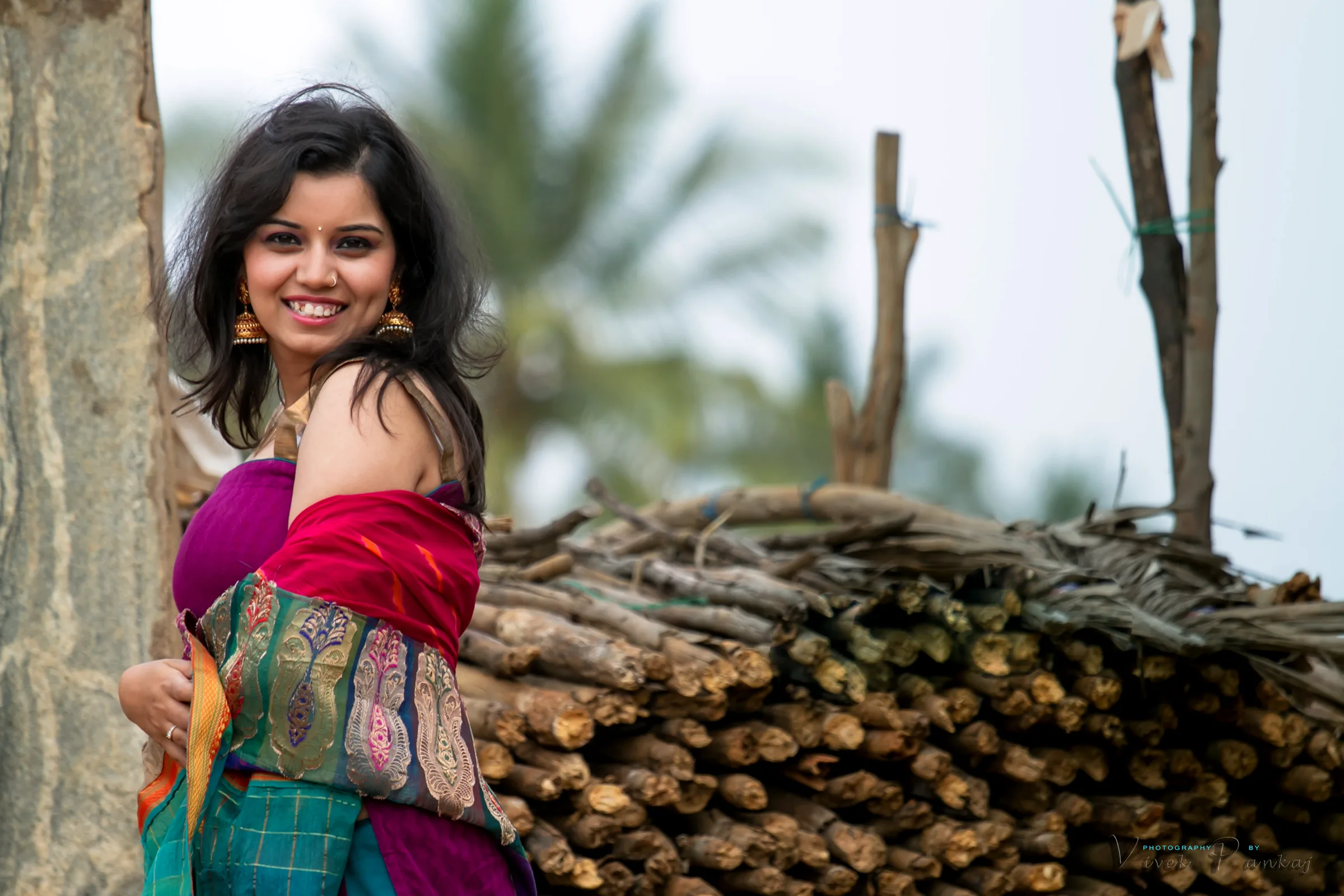 Smiling woman in a red shawl leaning on a stack of logs outdoors, from a pre-wedding shoot by Vivek Pankaj