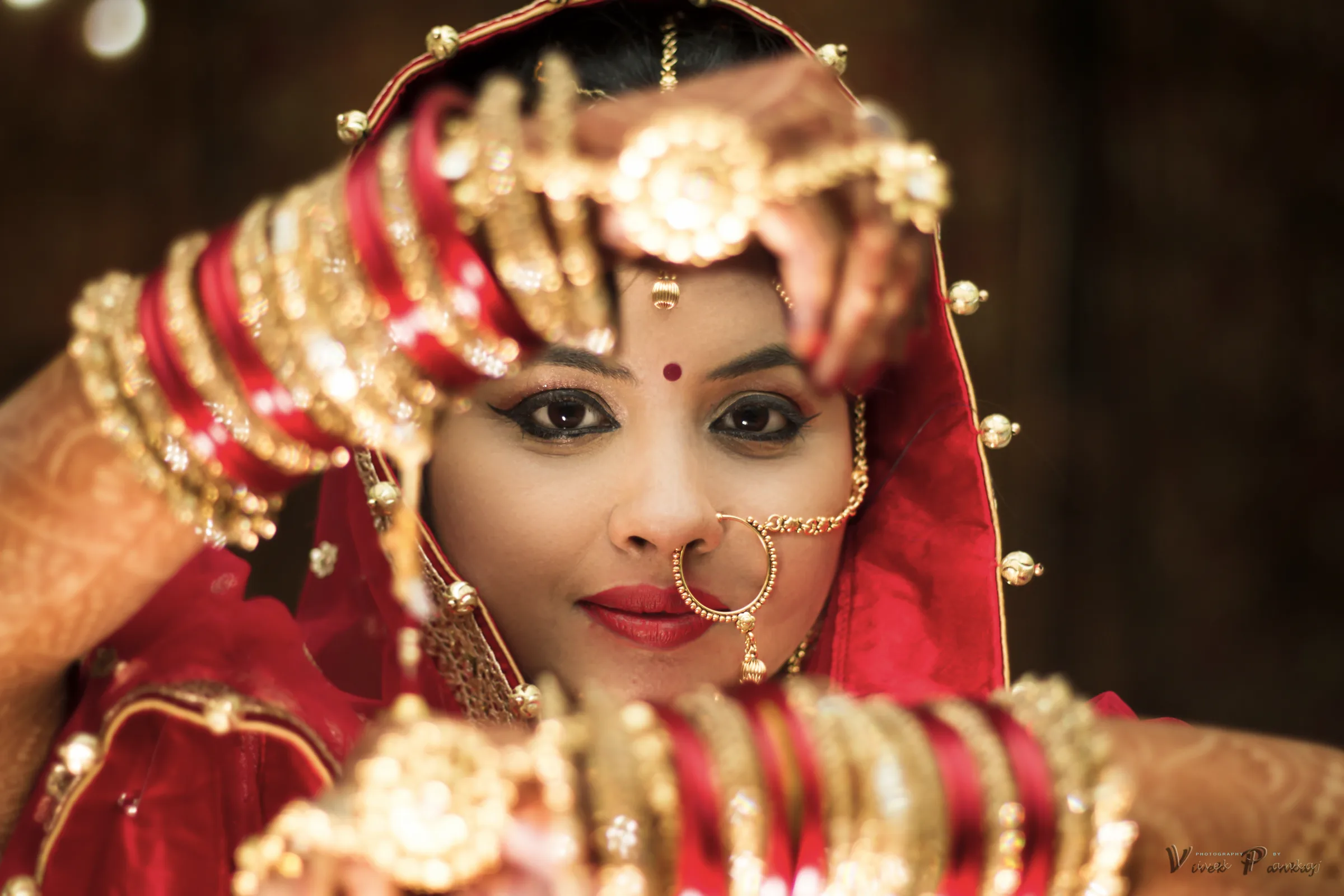 Bride in red with a gold nath and bangles framing her eyes with her hennaed hands, bridal portrait by Vivek Pankaj